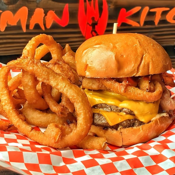 A double cheeseburger with bacon and onion rings, served on red and white checkered paper.