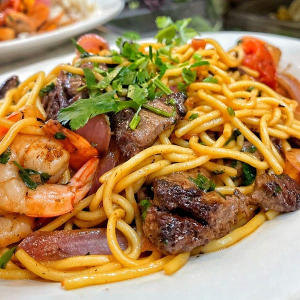 Peruvian Lomo Saltado with noodles, beef, shrimp, red onions, tomatoes, and cilantro on a white plate.